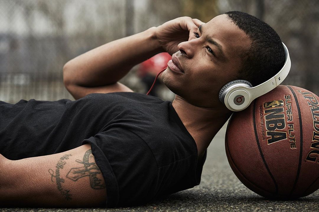 Young man in black shirt and headphones relaxes outdoors with a basketball, looking up and to the left, thoughtful expression.