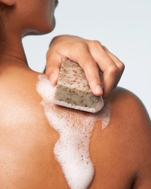Close-up of adult female washing her shoulder with a bar of soap and creating foam on skin in an indoor environment.