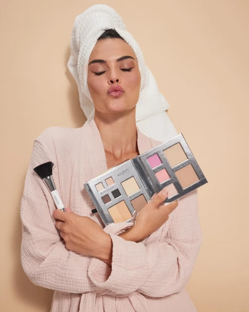 Woman in robe and towel poses with makeup palette and brush in a beauty shot, set against a neutral backdrop for beauty advertising.