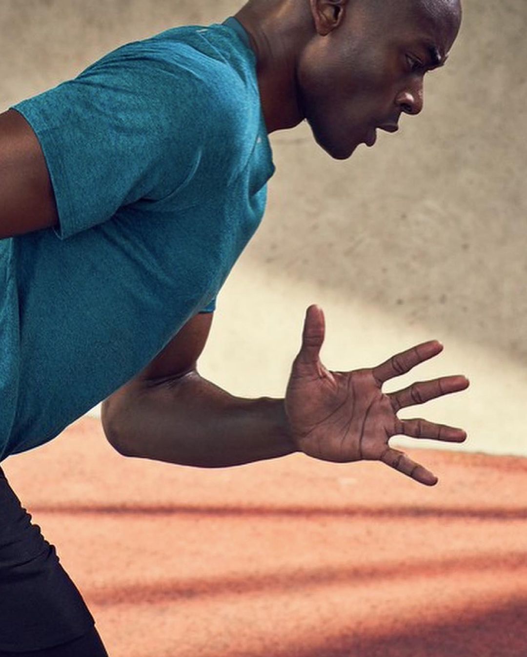 Athletic African-American man exercising outdoors on running track, wearing teal shirt, reaching forward with hand outstretched.