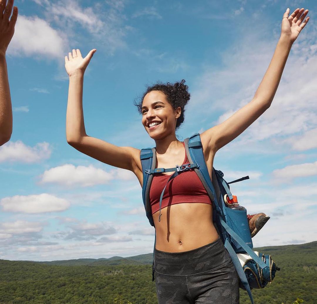 Woman with backpack raises her arms in celebration on a hike with a mountain vista under a bright sky with fluffy clouds.