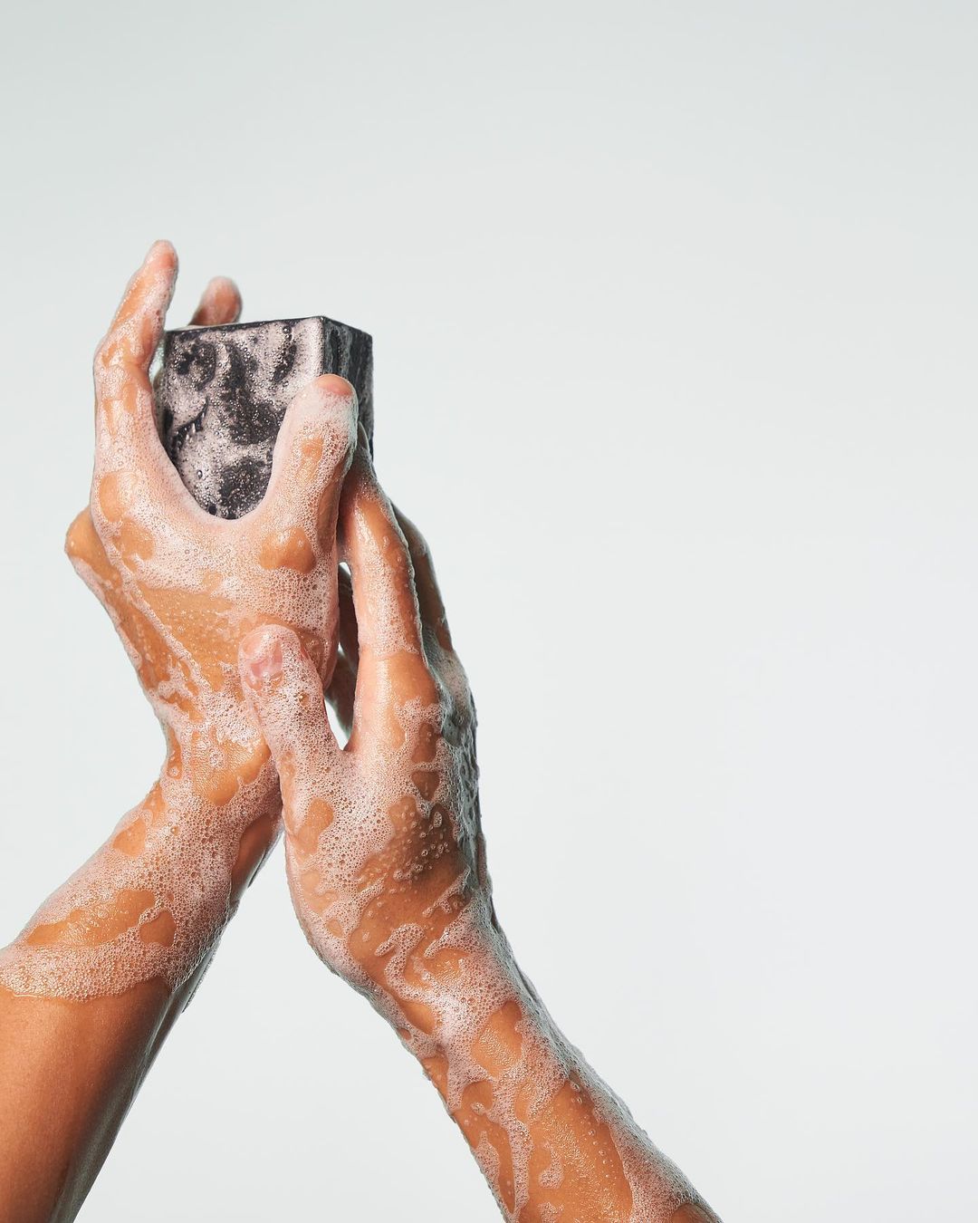 Close-up of two hands holding a dark-colored, rectangular bar of soap covered in white suds against a solid white backdrop.