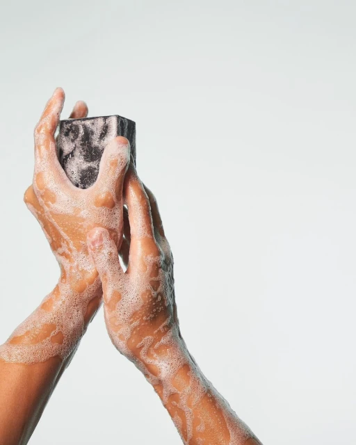 Close-up of two hands holding a dark-colored, rectangular bar of soap covered in white suds against a solid white backdrop.