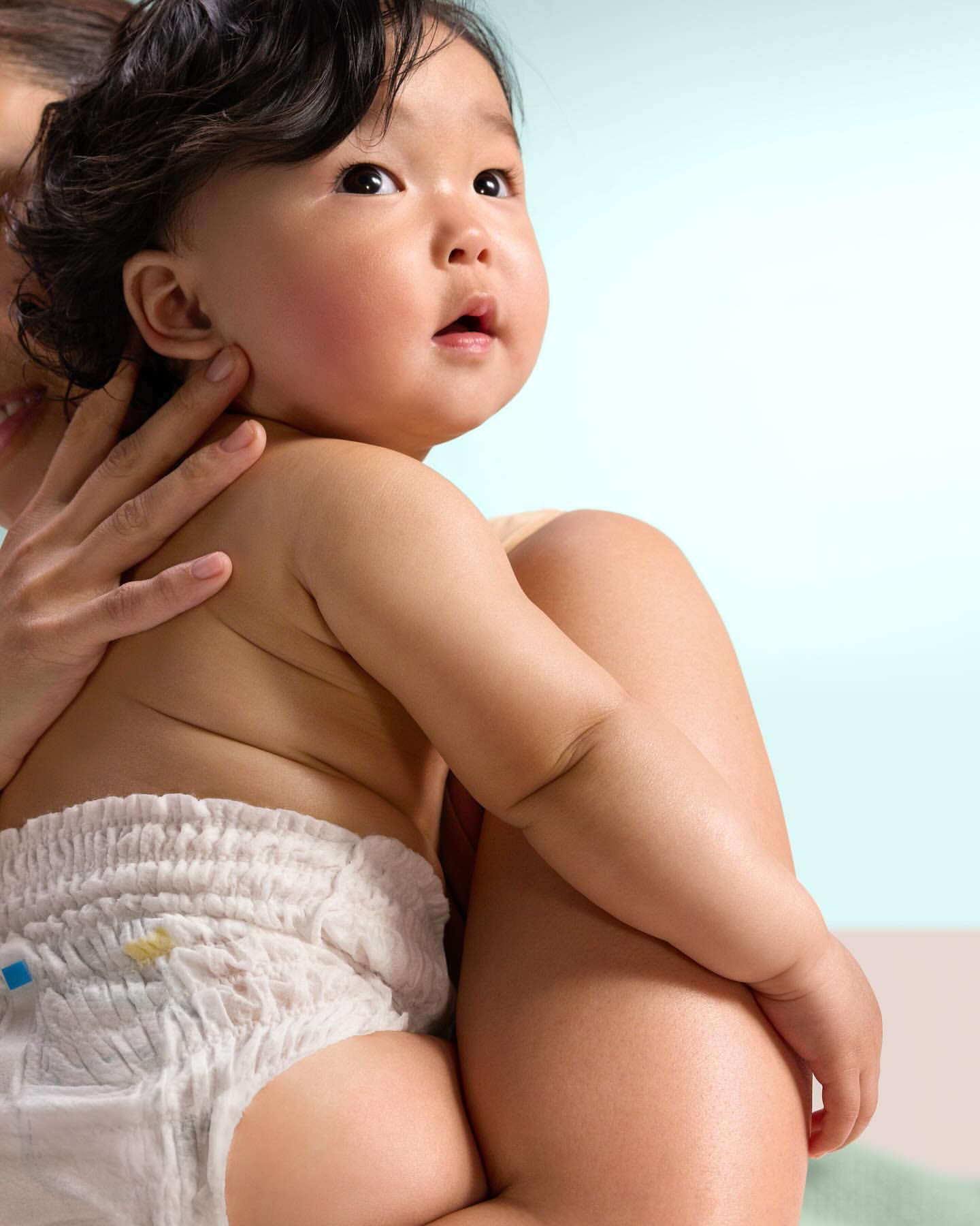 Cute Asian baby in a diaper being held by an adult indoors with a soft blue and white background. Child looking upward.