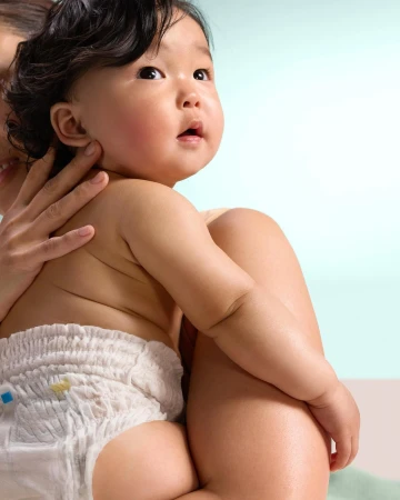 Cute Asian baby in a diaper being held by an adult indoors with a soft blue and white background. Child looking upward.