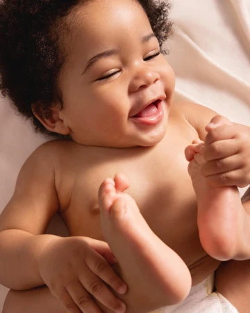 Close-up of a cheerful baby with dark curly hair, eyes closed, and a wide smile, lying on a cream-colored fabric surface in soft lighting.