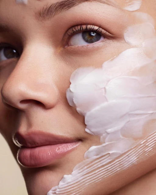 Close-up portrait of a woman's face with skincare cream applied. Light skin, light background, beauty shot, eye-level angle.