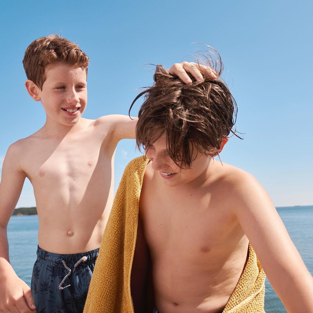 Two shirtless boys stand outdoors near water on a sunny day. One boy touches the other's hair as he holds a yellow towel.