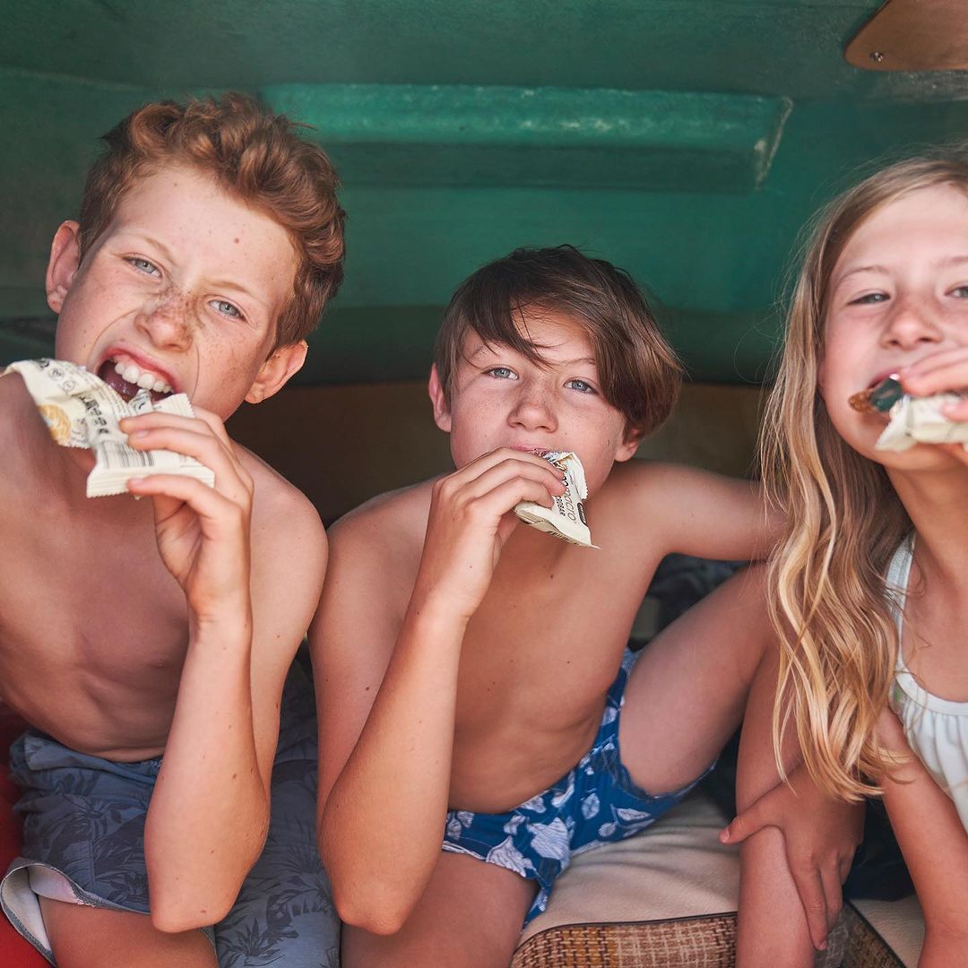 Three children, two boys and a girl, are eating snacks inside a structure with a green ceiling; summer, family, and childhood themes.