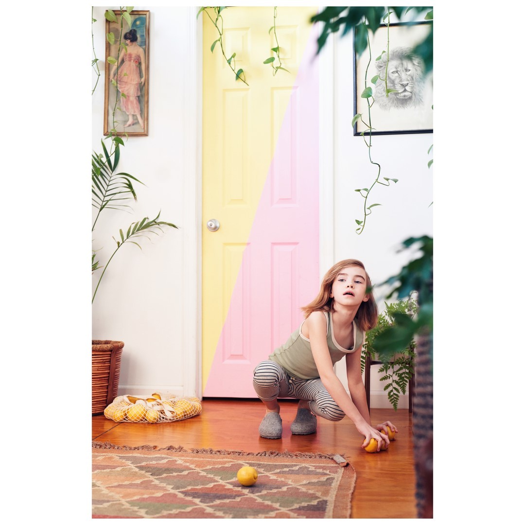 A young girl squats on a wooden floor, next to lemons and a colorful rug, with a pastel-colored door in the background, indoor setting.