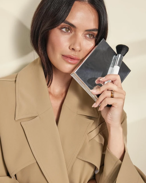 Brunette woman in trench coat poses with makeup brush and silver compact in a beauty shot with neutral backdrop.