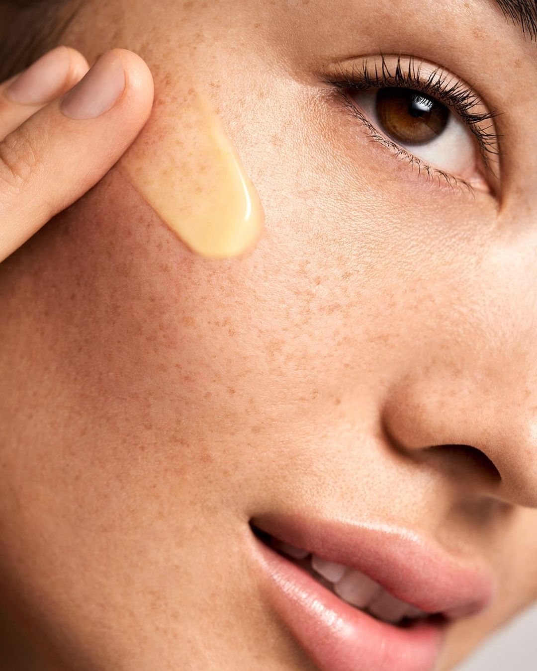 Close-up shot of a smiling woman with freckles applying a gold-colored serum to her face, highlighting skincare and beauty.