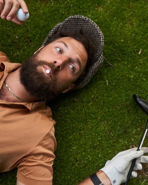 Man with a beard lying on green grass, wearing a cap and orange shirt, with a golf ball in his mouth and golf club in hand.