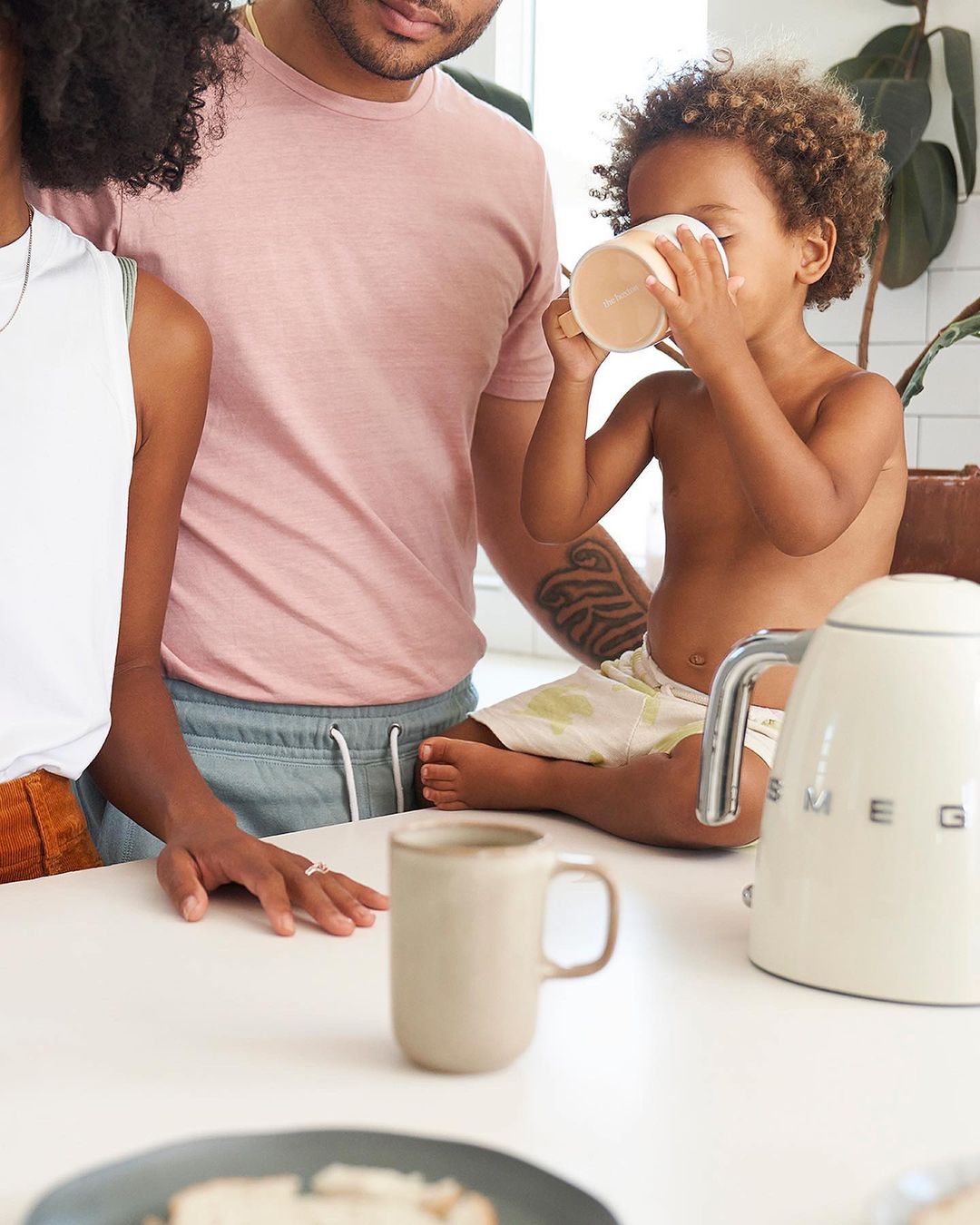 A family of three stands in a bright kitchen; a toddler drinks from a mug on the counter, while two adults stand nearby.