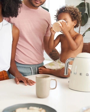 A family of three stands in a bright kitchen; a toddler drinks from a mug on the counter, while two adults stand nearby.