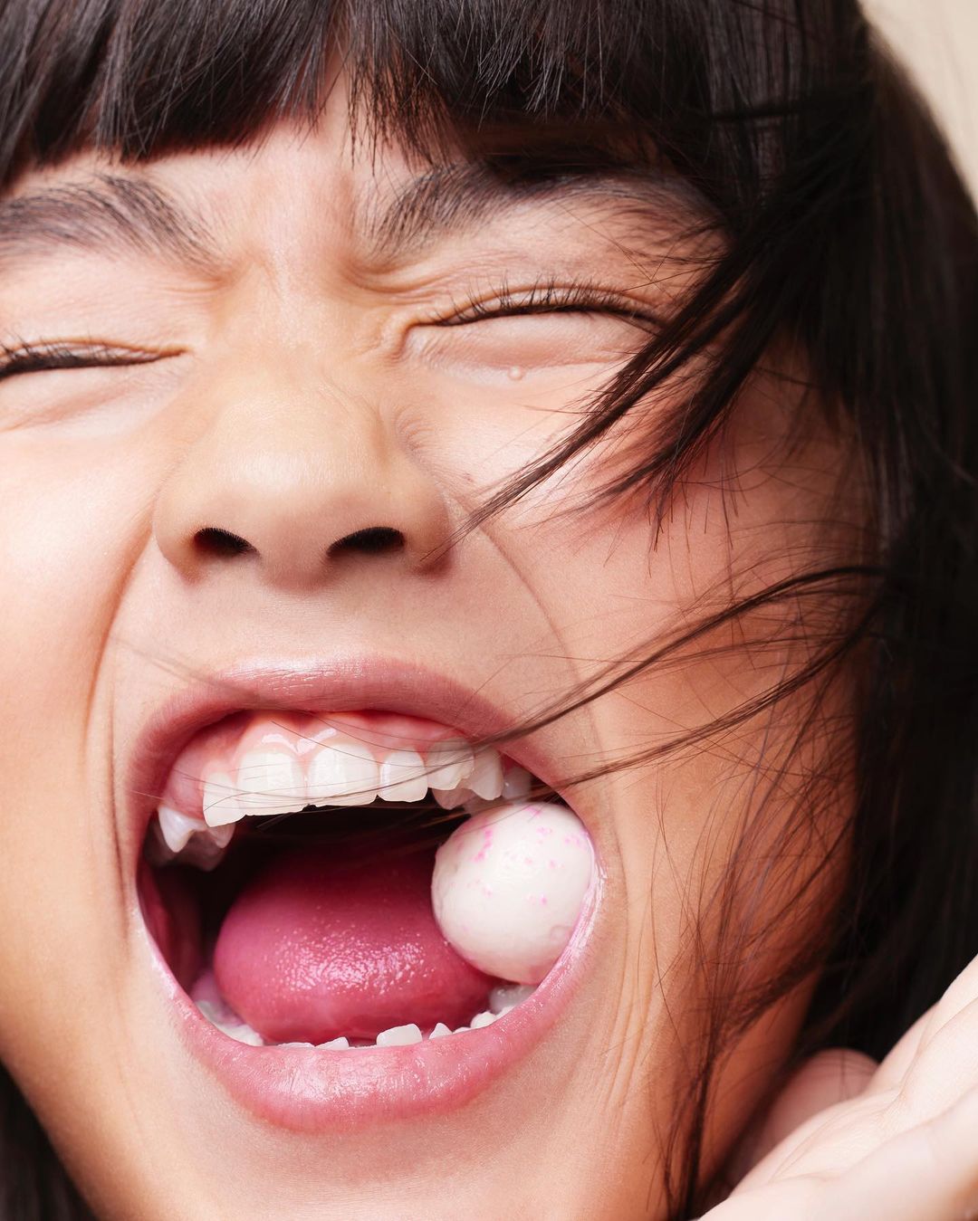 Close-up of a young girl with dark hair, eyes closed and mouth wide open, a round candy visible in her mouth, braces on her teeth.