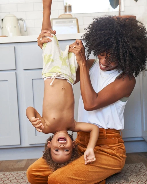 Mother holds her laughing toddler upside down in a bright, casual kitchen. Joyful family moment with light-colored cabinets.