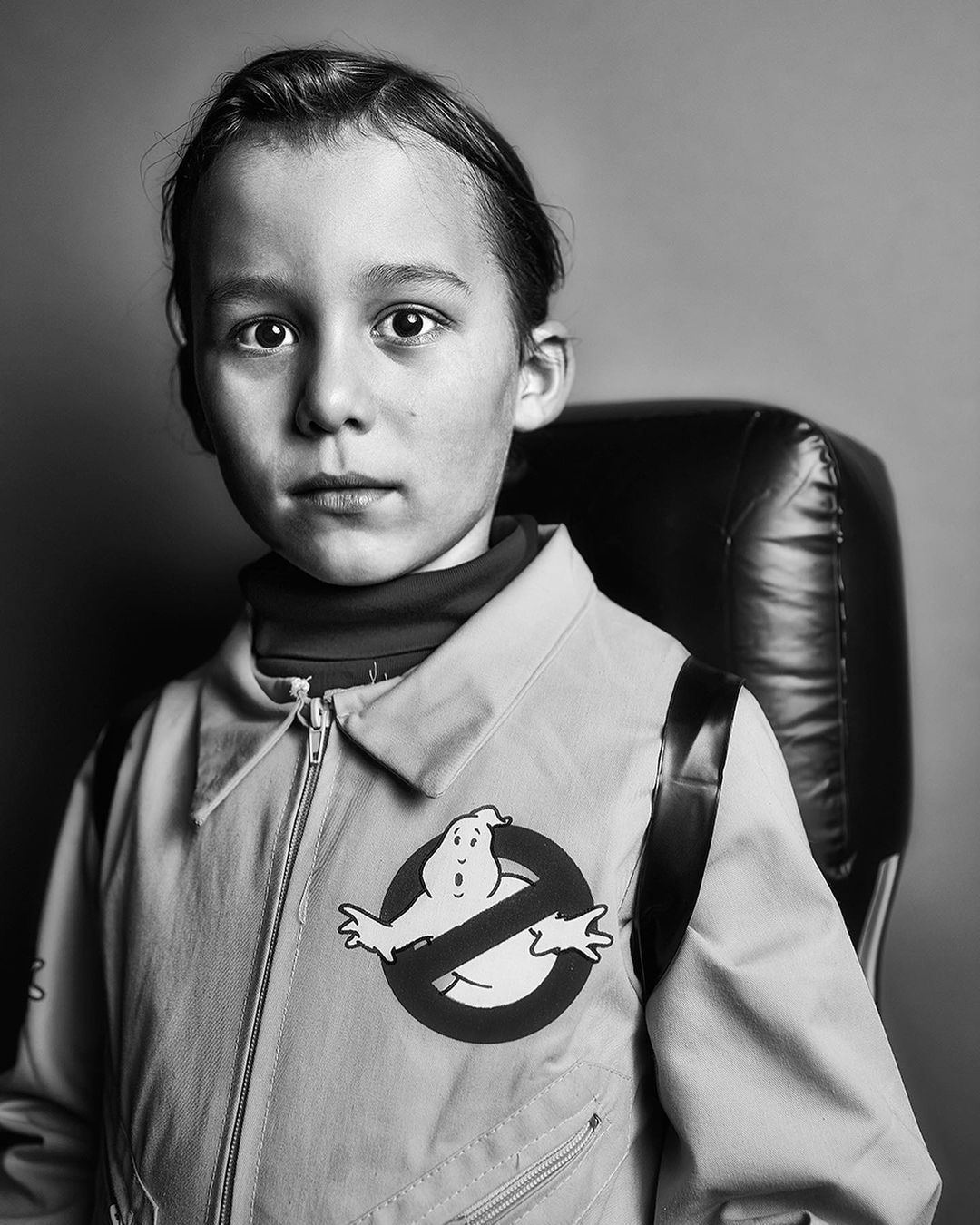 Portrait of a child wearing a Ghostbusters uniform, sitting in a leather chair, against a grey background. Costume detail.