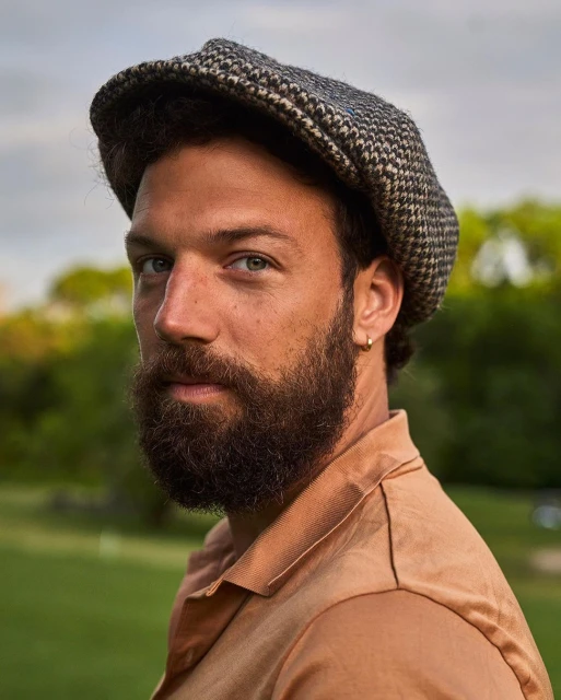 Portrait of a man with a beard wearing a tweed cap outdoors in a green setting. He has fair skin, green eyes, and a tan shirt.