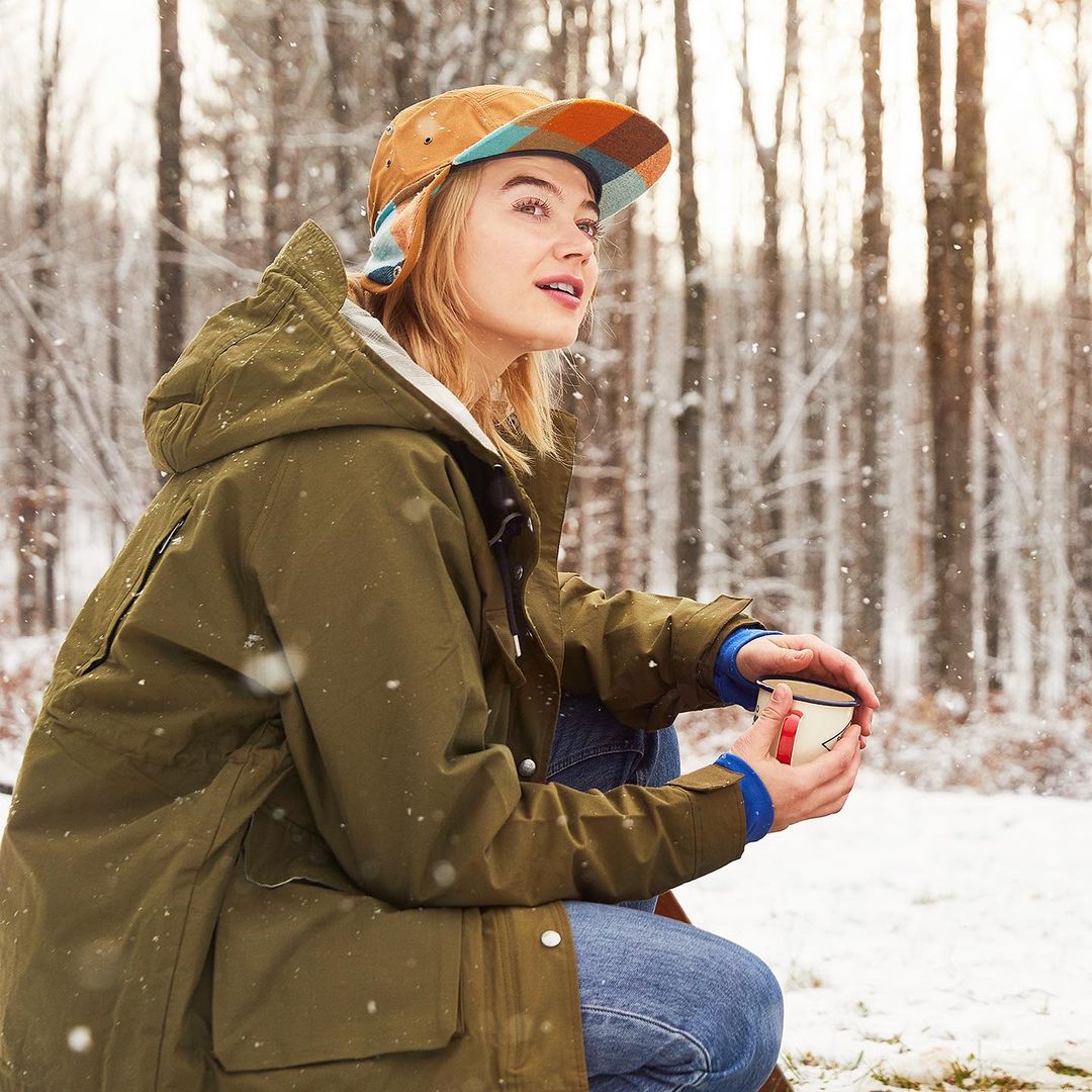 Adult female in olive parka and patterned cap crouches holding a mug, set against a snowy forest backdrop on an overcast day.
