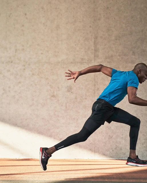 Athletic African-American man sprints on running track in blue shirt and black leggings, with cream wall background and horizontal lines.