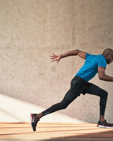 Athletic African-American man sprints on running track in blue shirt and black leggings, with cream wall background and horizontal lines.