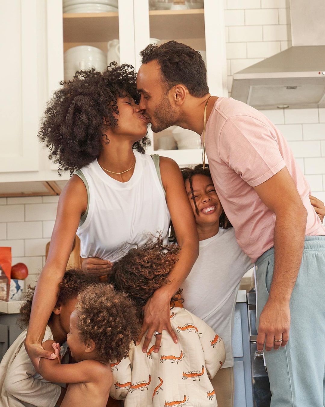 Family portrait in kitchen: parents kissing surrounded by four children, warm lighting, close-up framing, capturing a candid moment.