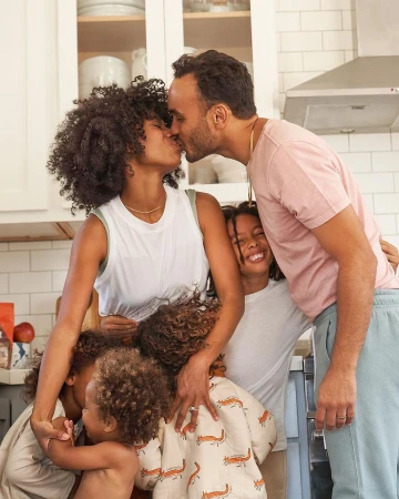 Family portrait in kitchen: parents kissing surrounded by four children, warm lighting, close-up framing, capturing a candid moment.