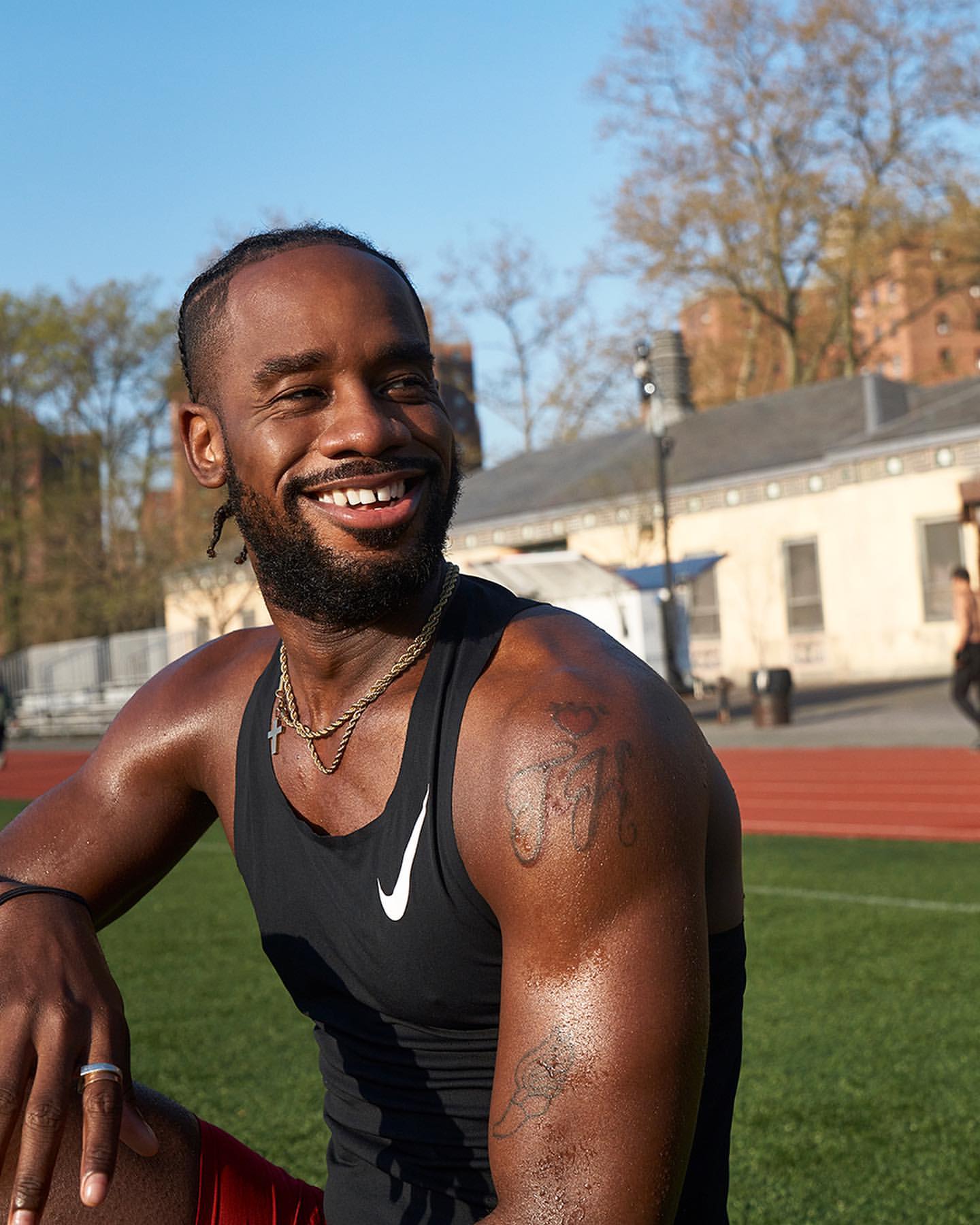 Smiling athlete on a track field wearing a tank top and gold chain on a sunny day. Building and trees in background.