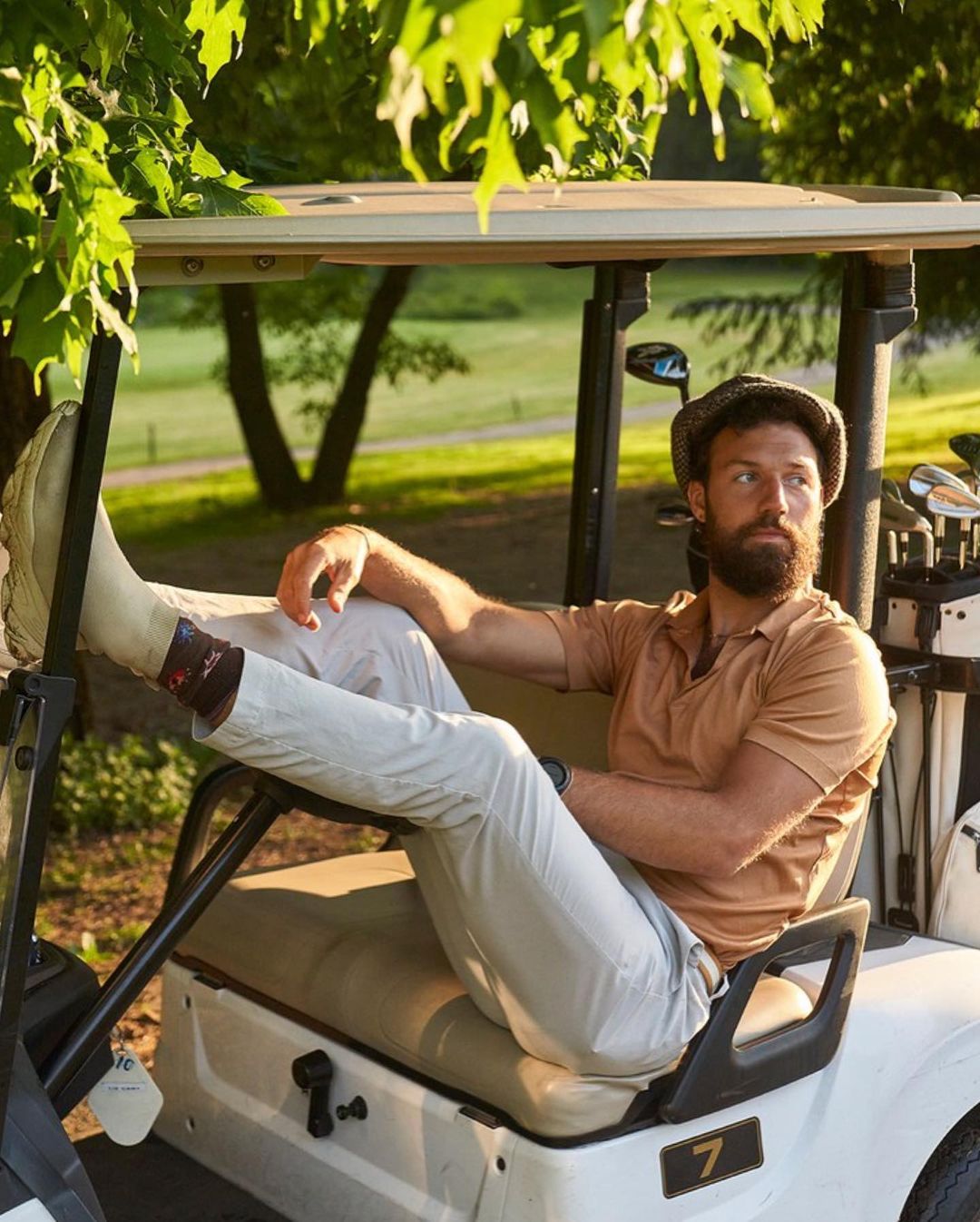 Adult male with a beard poses in a white golf cart on a golf course with trees in the background wearing casual attire.