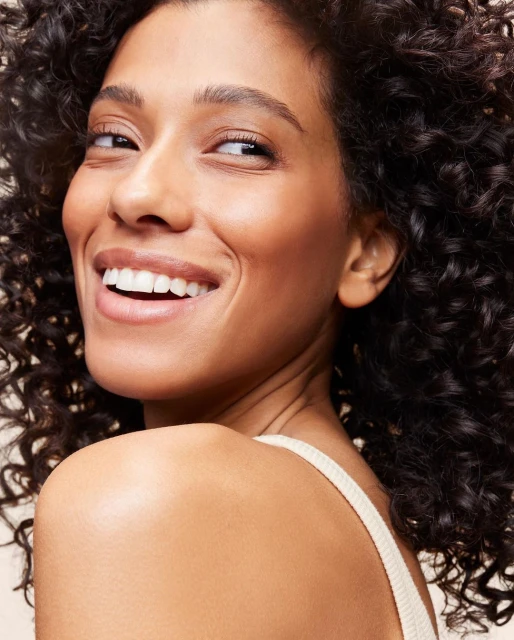 Close-up portrait of a smiling woman with curly dark hair, wearing a spaghetti-strap top, looking over her shoulder.
