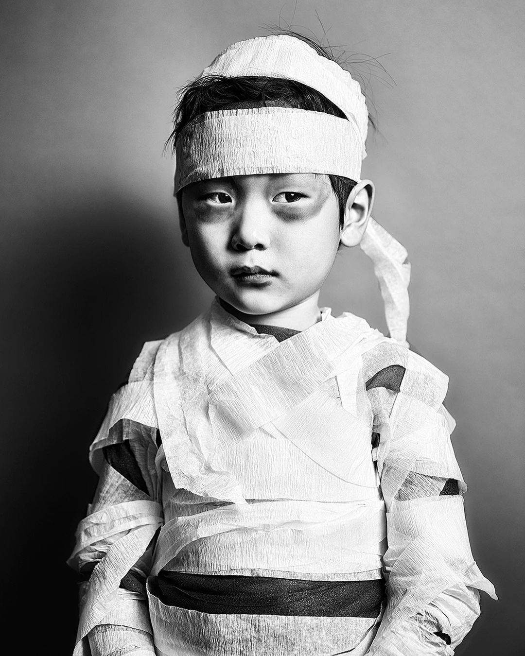 Black and white portrait of a young child dressed as a mummy with bandages wrapped around their head and torso against a grey background.