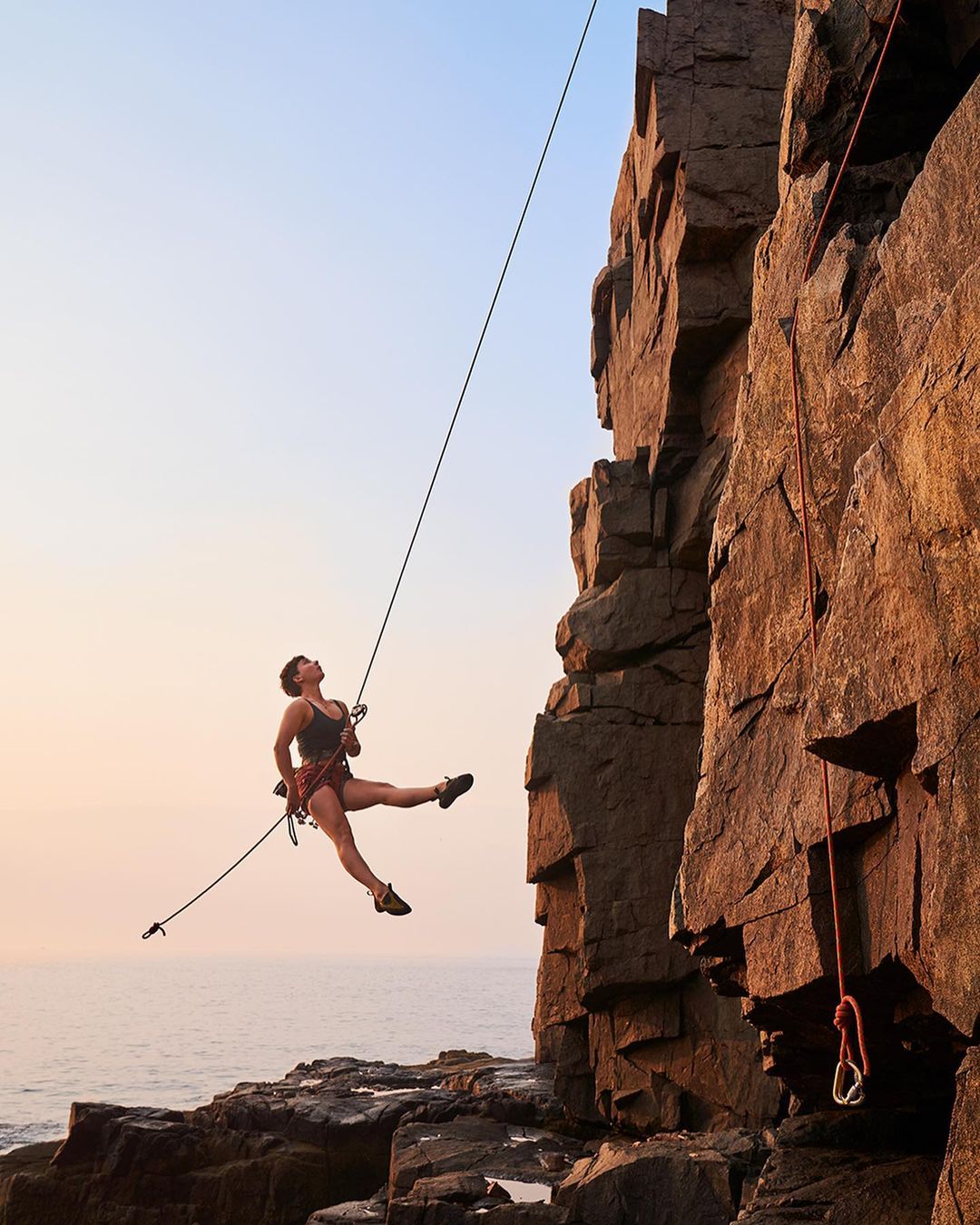 A woman in climbing gear suspended by ropes near a rock cliff face and the ocean, showcasing outdoor fitness and rock climbing.