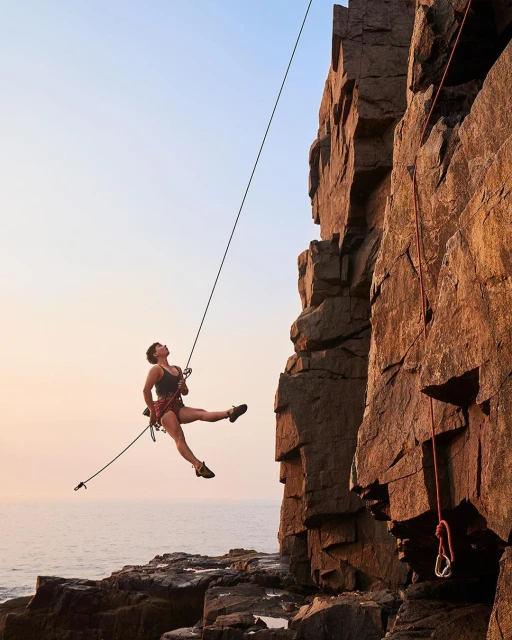 A woman in climbing gear suspended by ropes near a rock cliff face and the ocean, showcasing outdoor fitness and rock climbing.