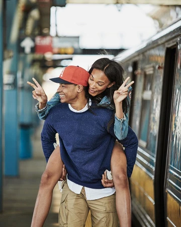 Smiling couple in casual clothing poses at train station; man gives woman a piggyback ride while both flash the peace sign.