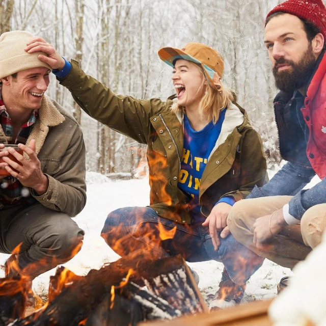 Three friends laughing near a campfire in the snowy woods. One man touches another man's head playfully while the other man smiles.