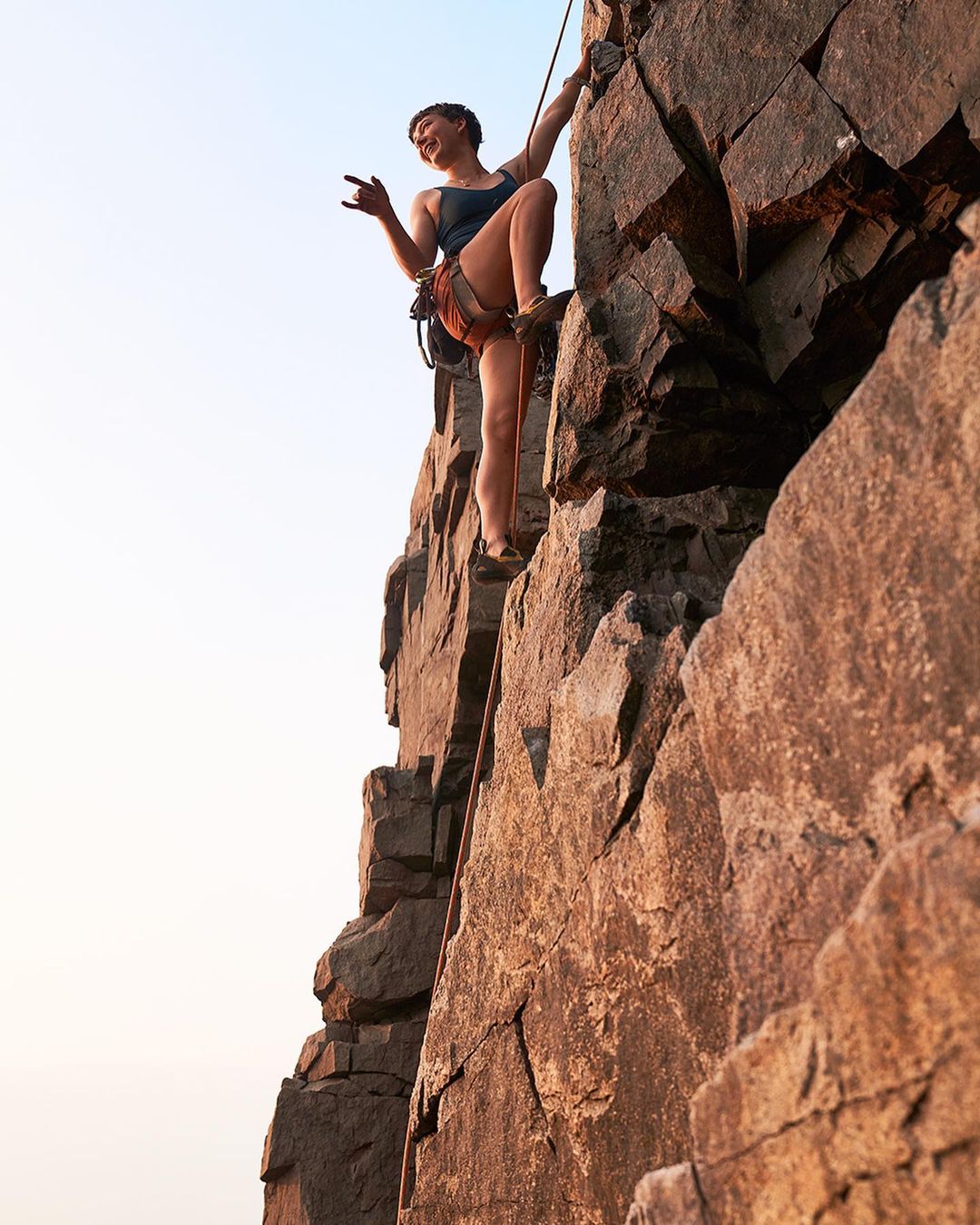Female rock climber ascends a jagged rock face outdoors, using ropes and safety gear with a clear sky in the background.