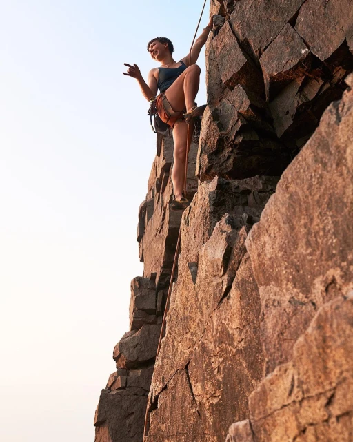 Female rock climber ascends a jagged rock face outdoors, using ropes and safety gear with a clear sky in the background.