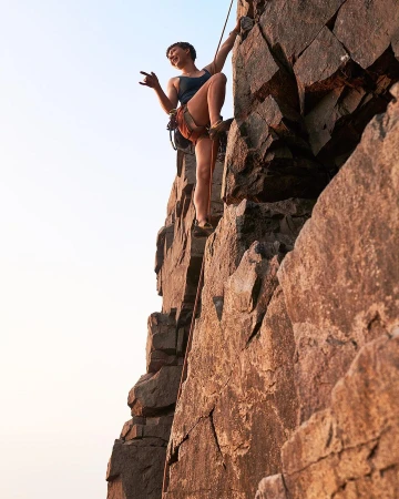 Female rock climber ascends a jagged rock face outdoors, using ropes and safety gear with a clear sky in the background.