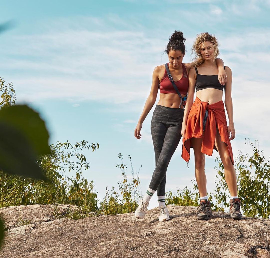 Two adult women in athletic wear pose on a rocky hilltop with a scenic sky backdrop, showcasing active lifestyle and outdoor fitness.