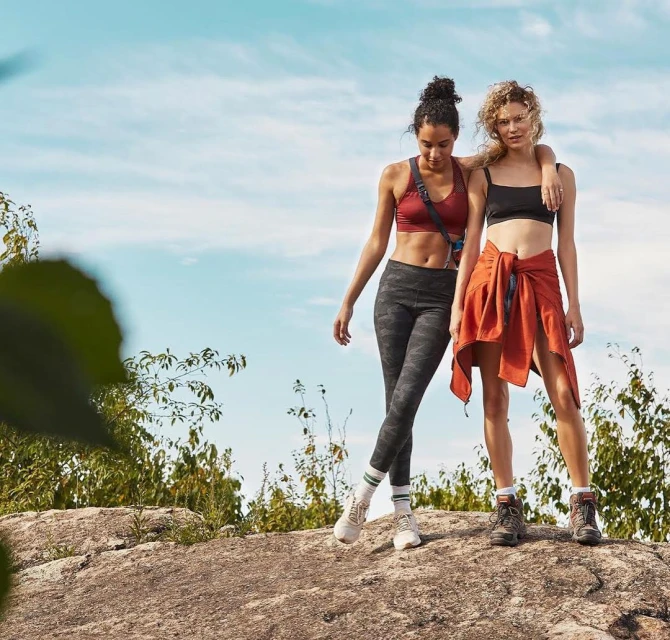 Two adult women in athletic wear pose on a rocky hilltop with a scenic sky backdrop, showcasing active lifestyle and outdoor fitness.