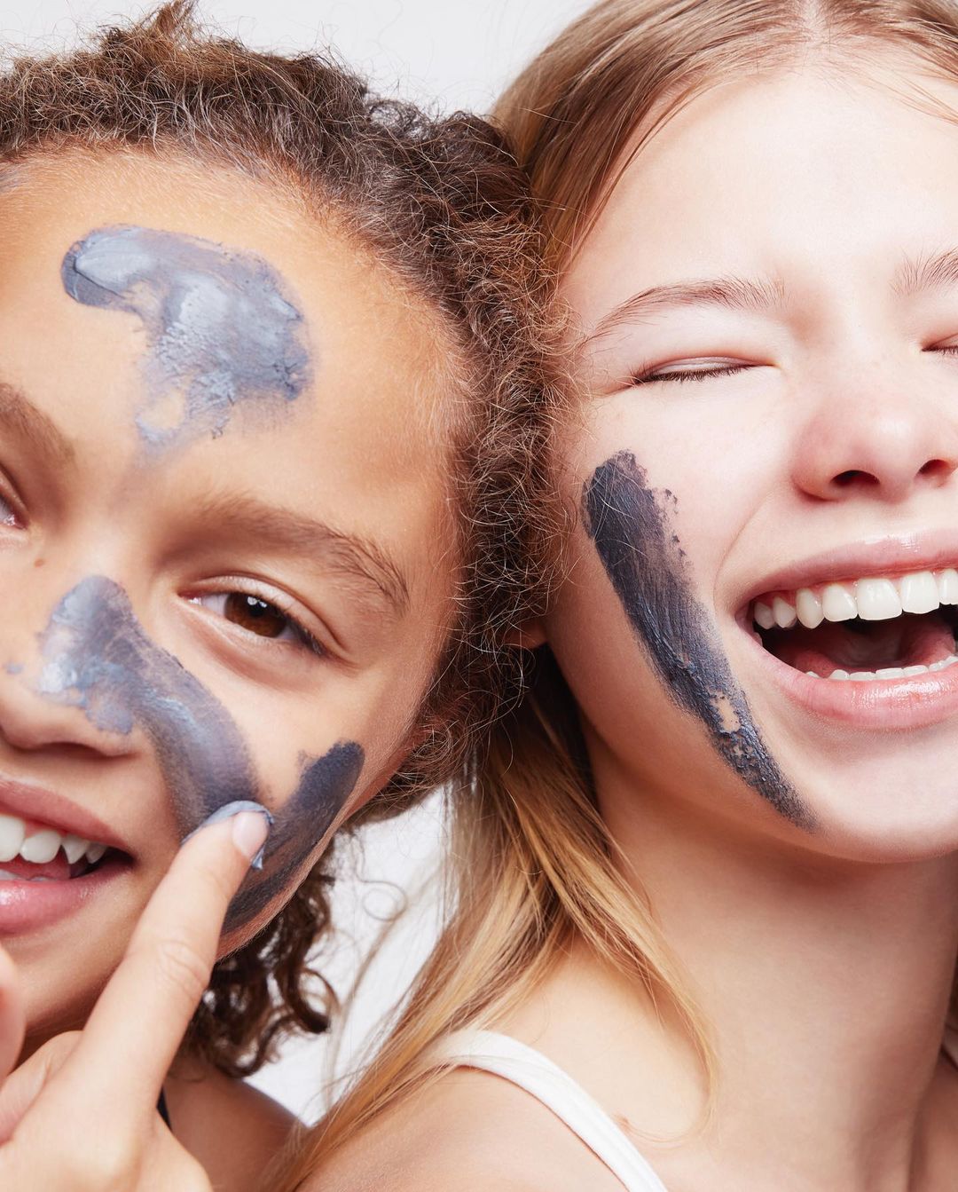 Two young women apply a grey clay mask to their faces. Both have big smiles and look directly into the camera in a close-up shot.
