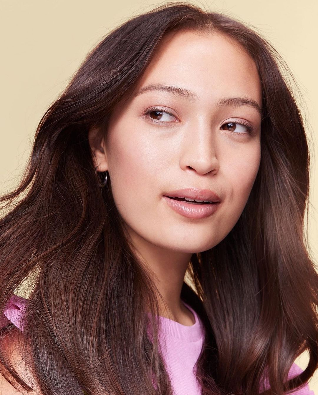 Headshot of a young woman with long brunette hair wearing a pink top in a bright studio setting. Beauty and fashion concept.