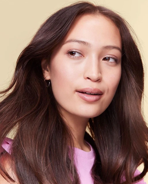 Headshot of a young woman with long brunette hair wearing a pink top in a bright studio setting. Beauty and fashion concept.