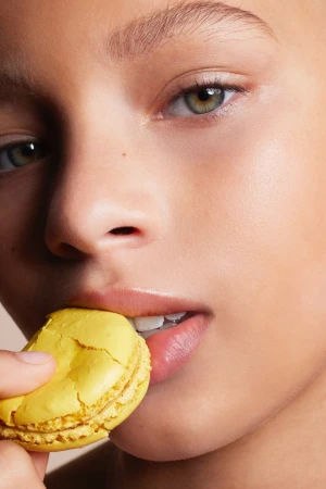 Close-up of a fair-skinned young adult with green eyes, biting a yellow macaroon in a studio environment with soft lighting.