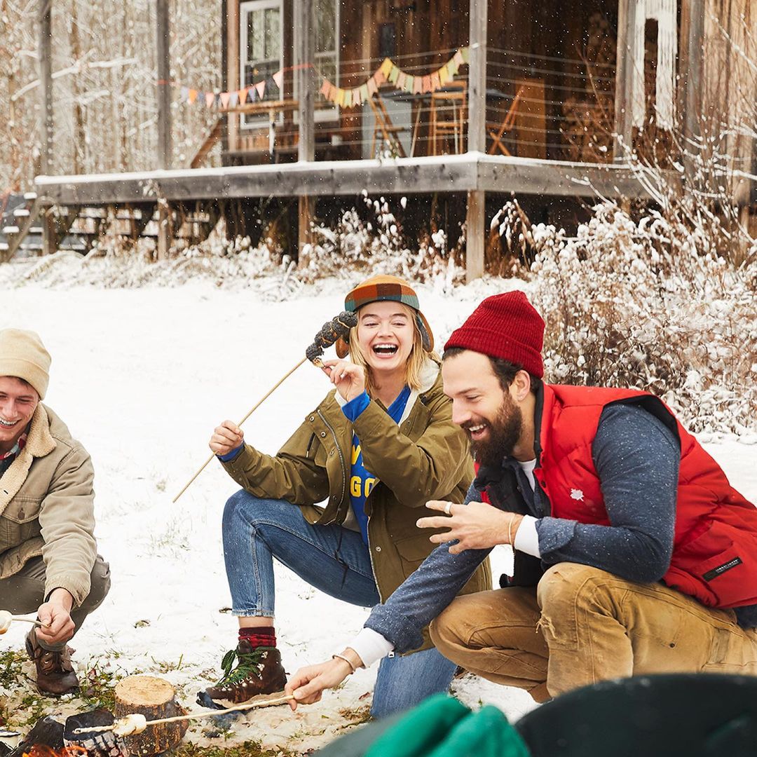 Three people smile while roasting marshmallows over a fire pit in a snowy forest setting with a cabin in the background.