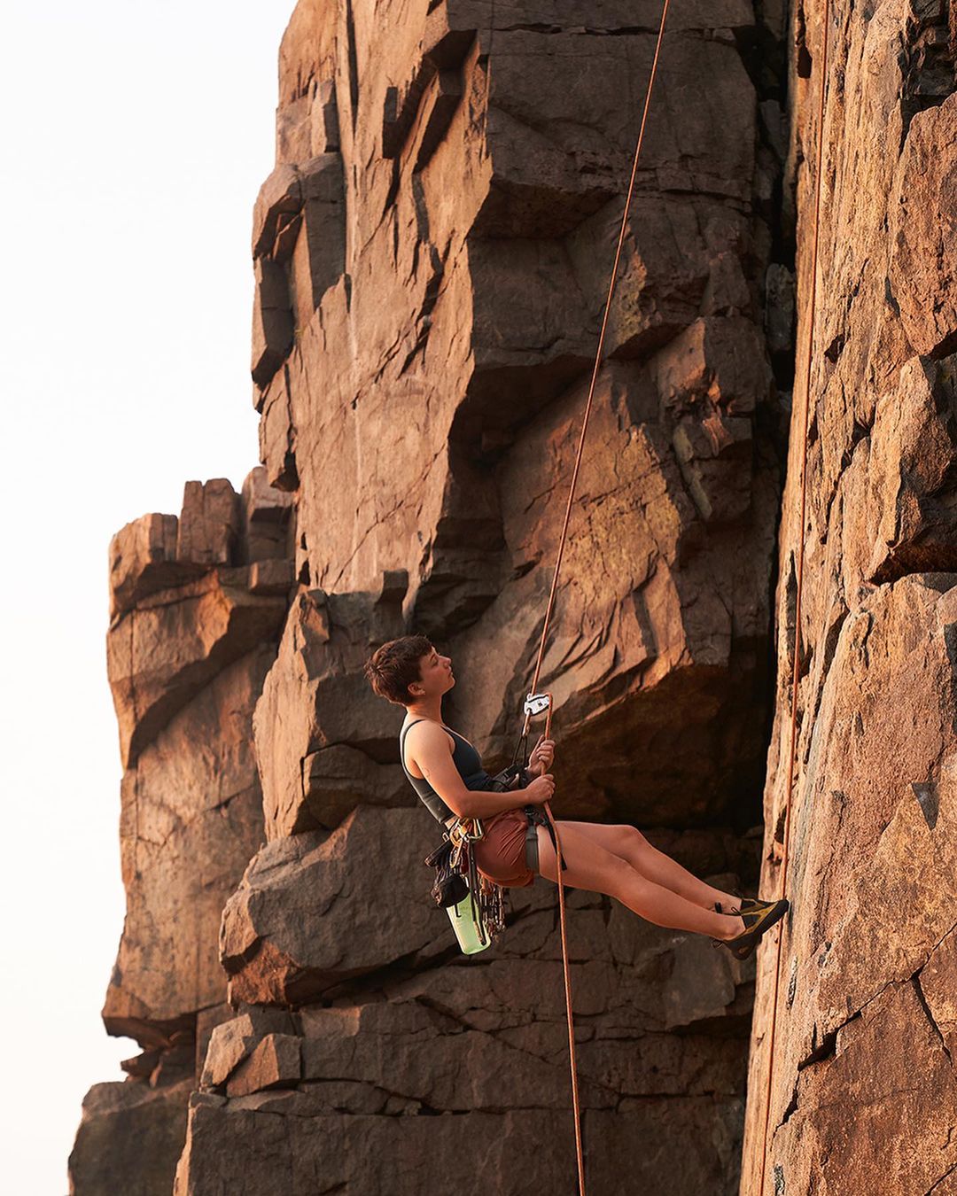 A woman rock climbing on a sheer cliff face, suspended by a rope, wearing athletic shorts and a tank top, with climbing gear visible.