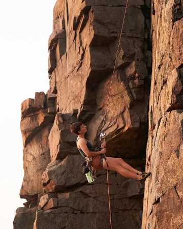 A woman rock climbing on a sheer cliff face, suspended by a rope, wearing athletic shorts and a tank top, with climbing gear visible.