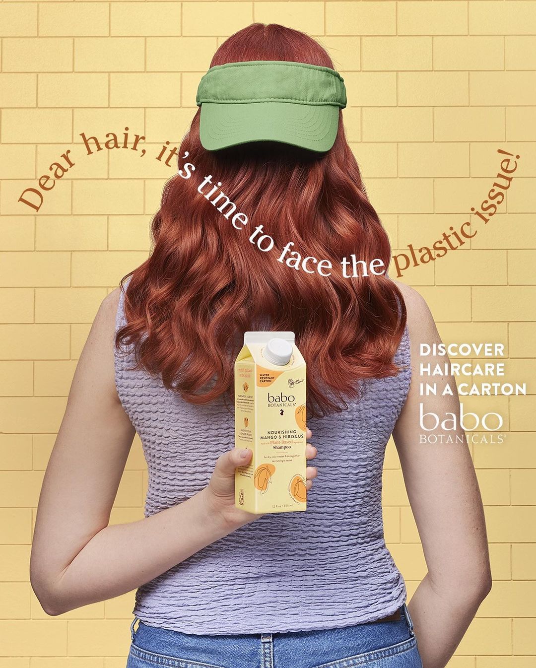 Back view of a redhead adult female wearing a green visor, holding a carton of Babo Botanicals haircare product in a yellow setting.