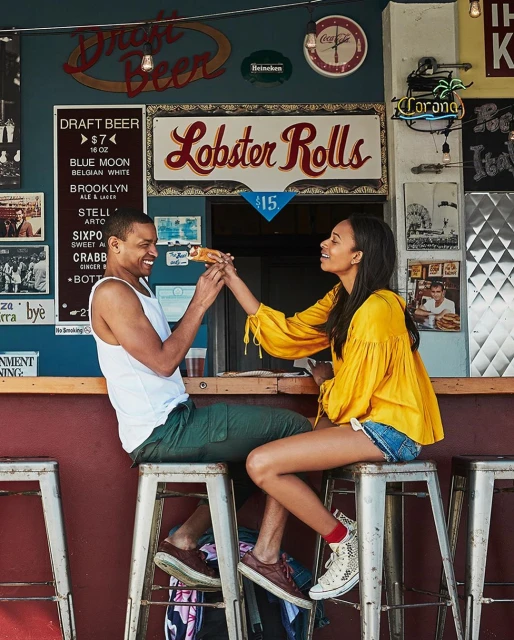 A man and woman sharing a lobster roll at an outdoor bar with vintage signs. The woman is handing the sandwich to the smiling man.